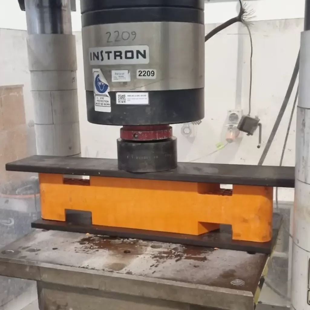 A hydraulic press labeled "Instron" compresses a steel I-beam resting on an orange support structure in a laboratory setting.