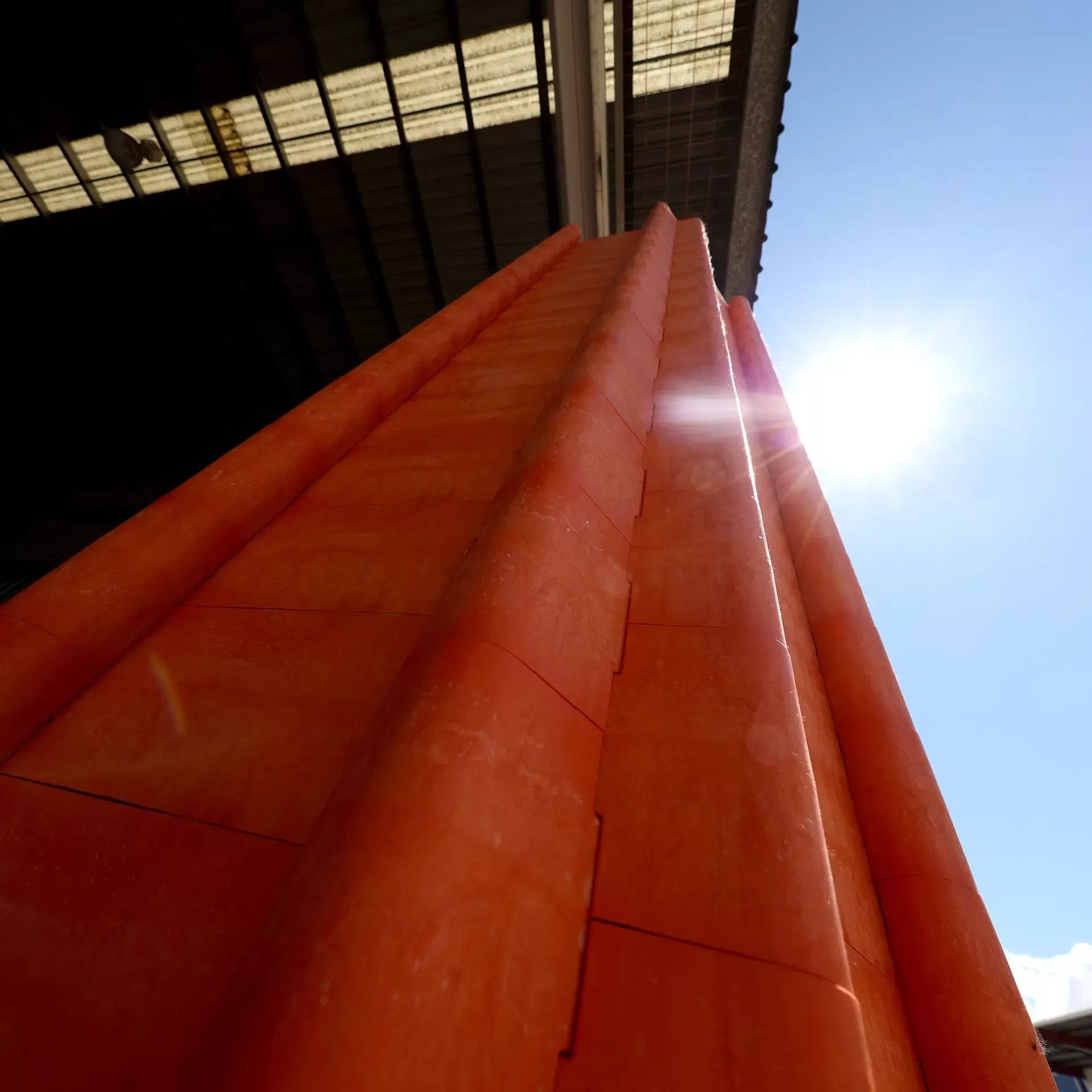 Large orange pipes are stacked vertically under a metal roof, with sunlight shining brightly in the background.